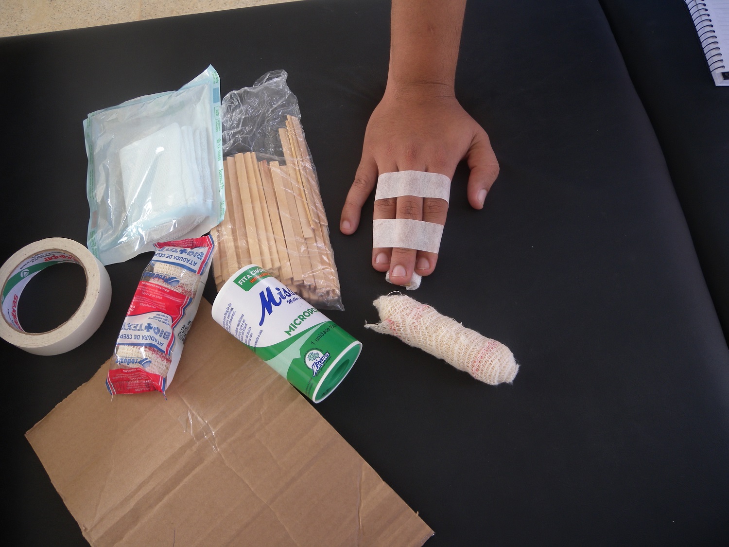 Demonstração de como fazer imobilização de dedo fraturado com materiais como palito de picolé, fita crepe, durex, papelão